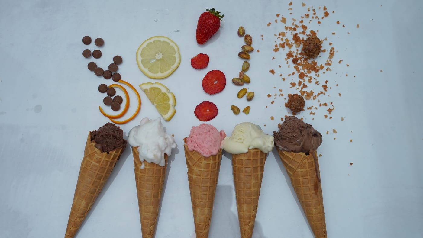 Five gelato cones with their ingredients laid out above them on a light ground