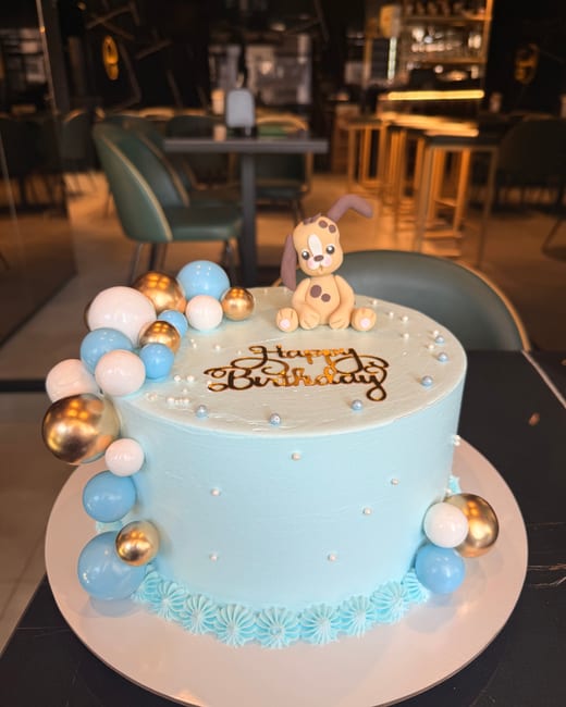 A pale blue cake with a puppy figure and a garland of chocolate spheres, on a table at the patisserie